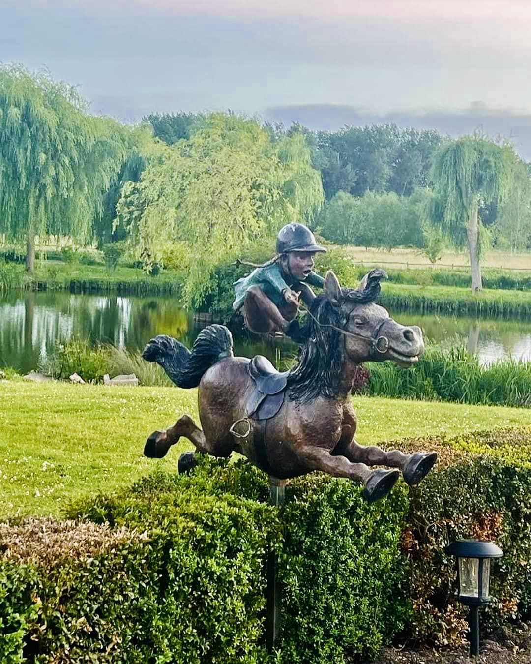 A metal statue depicting a horse and rider gracefully leaping over a hedge, showcasing dynamic movement and artistry.